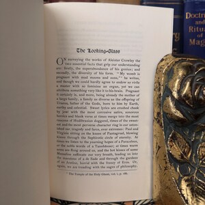 Pu&ograve; includere: Un primo piano di una pagina di libro con il titolo "The Looking-Glass" e testo sulle opere di Aleister Crowley. Il testo tratta della diversit&agrave; del suo genio e dell'abbondanza della sua forma. La pagina &egrave; ingiallita dal tempo e ha un aspetto usurato e vintage.