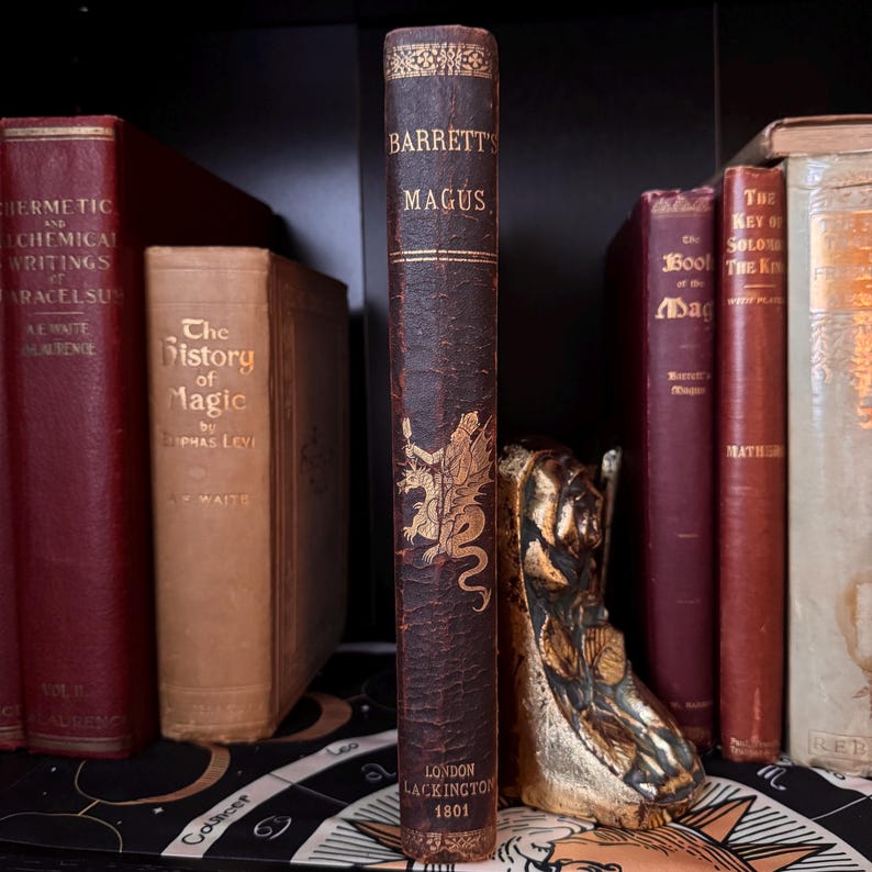 Puede incluir: Una estanter&iacute;a con libros antiguos. El libro central, "Barrett's Magus", es marr&oacute;n con letras doradas y un dise&ntilde;o de drag&oacute;n. Otros libros son rojos, beige y crema. Tambi&eacute;n se ve una estatua dorada.