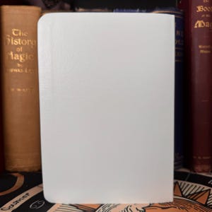 May include: A white, blank journal with a textured cover stands upright, its pages ready for writing. The journal is positioned in front of a collection of antique books with gold lettering and dark red covers, suggesting a theme of history or magic.