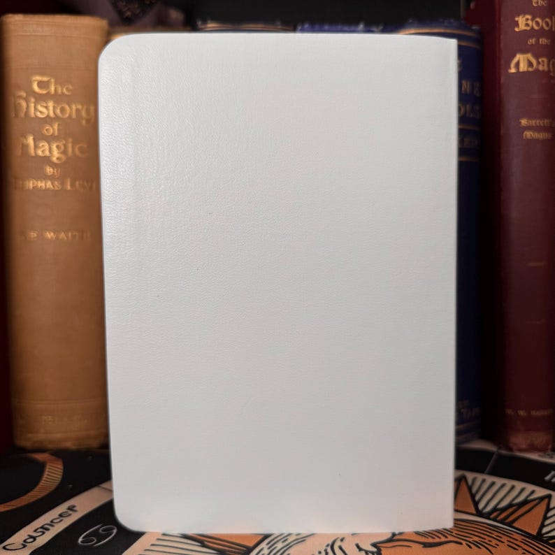 May include: A white, blank journal with a textured cover stands upright, its pages ready for writing. The journal is positioned in front of a collection of antique books with gold lettering and dark red covers, suggesting a theme of history or magic.