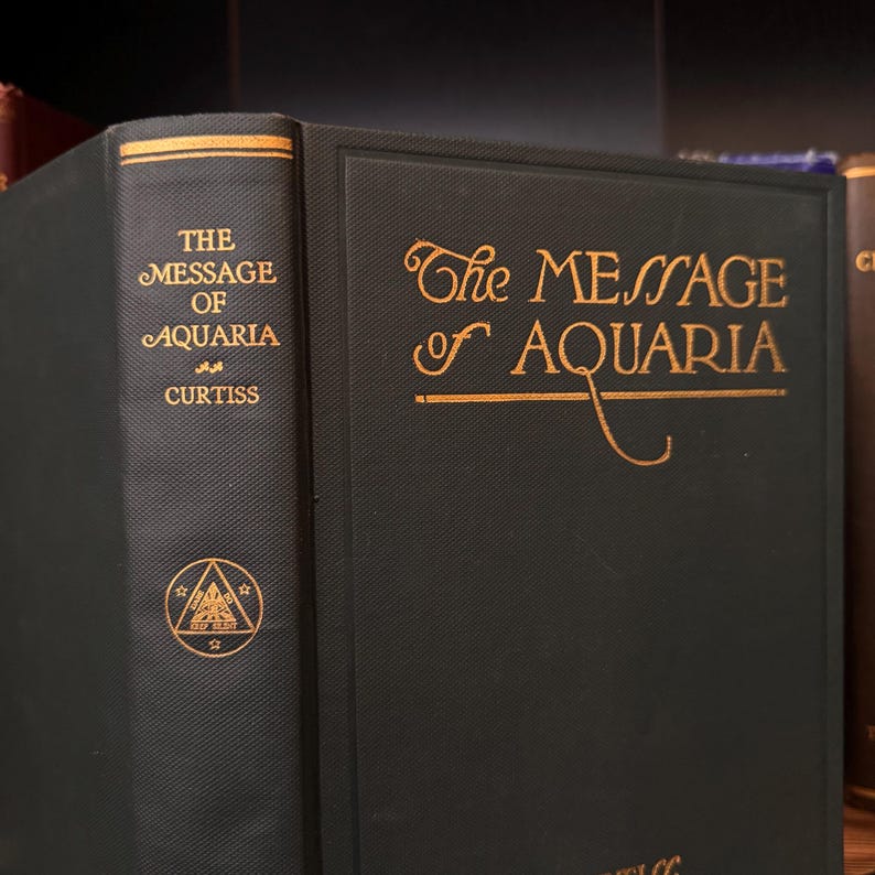 May include: A dark green hardcover book with gold lettering on the spine and front cover. The title is "The Message of Aquaria" by Curtiss. The front cover also features a gold symbol of a triangle with a star at each point.