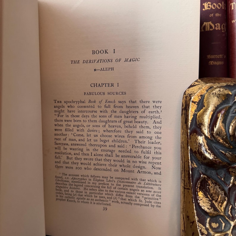 Puede incluir: Un libro antiguo titulado "Book I The Derivations of Magic" con un cap&iacute;tulo titulado "Fabulous Sources". El texto trata sobre el Libro de Enoc ap&oacute;crifo y la historia de los &aacute;ngeles que cayeron del cielo para tener hijos con las hijas de la tierra. El libro est&aacute; abierto en la p&aacute;gina 39.
