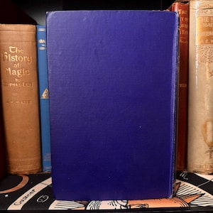 May include: A selection of vintage books with diverse colours and textures. The spine of a dark blue book is central, with other books in shades of brown, red, and cream in the background. The books are arranged on a shelf.