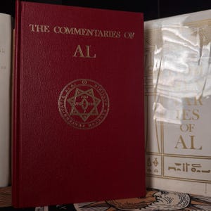 May include: A burgundy book titled "The Commentaries of AL" with gold lettering and a gold emblem. The book is bound in a textured material. Another book is partially visible to the right, with white and gold text.
