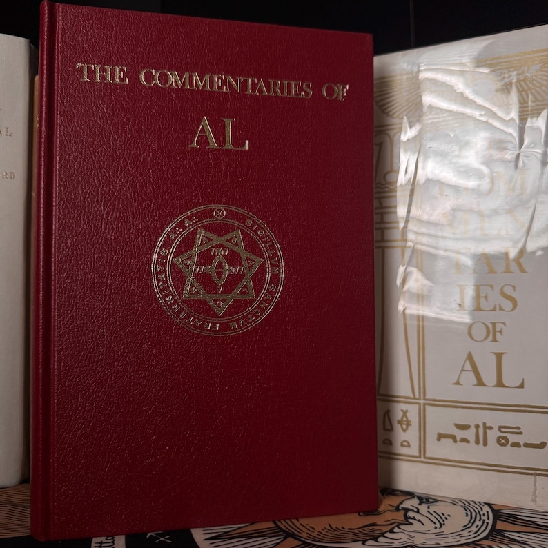 May include: A burgundy book titled "The Commentaries of AL" with gold lettering and a gold emblem. The book is bound in a textured material. Another book is partially visible to the right, with white and gold text.