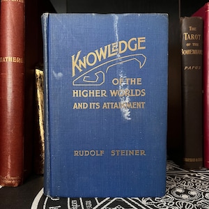 May include: A blue hardcover book titled "Knowledge of the Higher Worlds and Its Attainment" by Rudolf Steiner. The book cover features a gold illustration of a stylized archway.