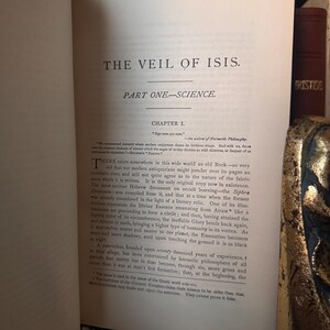 May include: Open book with the title "THE VEIL OF ISIS" and the subtitle "PART ONE-SCIENCE." The text is in black ink on aged, off-white paper. The book is open to Chapter 1, with detailed text and annotations. The book's spine and a decorative gold element are visible.