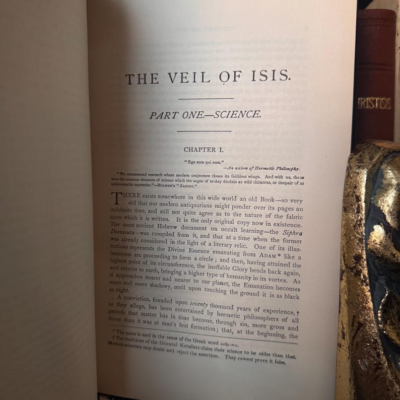 May include: Open book with the title "THE VEIL OF ISIS" and the subtitle "PART ONE-SCIENCE." The text is in black ink on aged, off-white paper. The book is open to Chapter 1, with detailed text and annotations. The book's spine and a decorative gold element are visible.