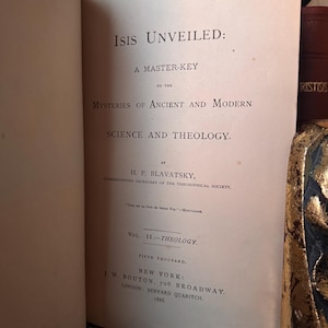 May include: Open book titled "Isis Unveiled" by H.P. Blavatsky. The book's title and author are prominently displayed in black text on a cream-colored page. The book is a master key to the mysteries of ancient and modern science and theology.