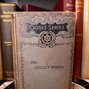 Pu&ograve; includere: Un libro vintage intitolato "The Occult World" della "Occult Series" con una stella dorata e un simbolo di croce sulla copertina.