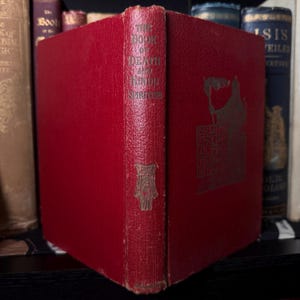 May include: A vintage book with a red cover titled "The Book of Death and Hindu Spiritism." The spine and cover feature gold lettering and decorative elements. The book is displayed among other antique books on a shelf.