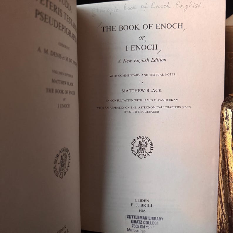 May include: Open book titled "The Book of Enoch, or 1 Enoch" by Matthew Black. The book is a new English edition with commentary and textual notes. The cover is off-white with black text. The book was published in Leiden in 1985.
