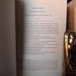 May include: Open book with text titled "The Book of Enoch" and "The First Vision of Enoch." The pages are yellowed, with text in black. A decorative gold object is visible on the right side of the image.