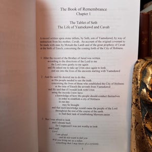 May include: Open book page with text titled "The Book of Remembrance, Chapter 1". The text describes "The Tablet of Seth" and "The Life of Yaatsekawd and Cavah". The page is white with black text. A decorative gold object is on the right.