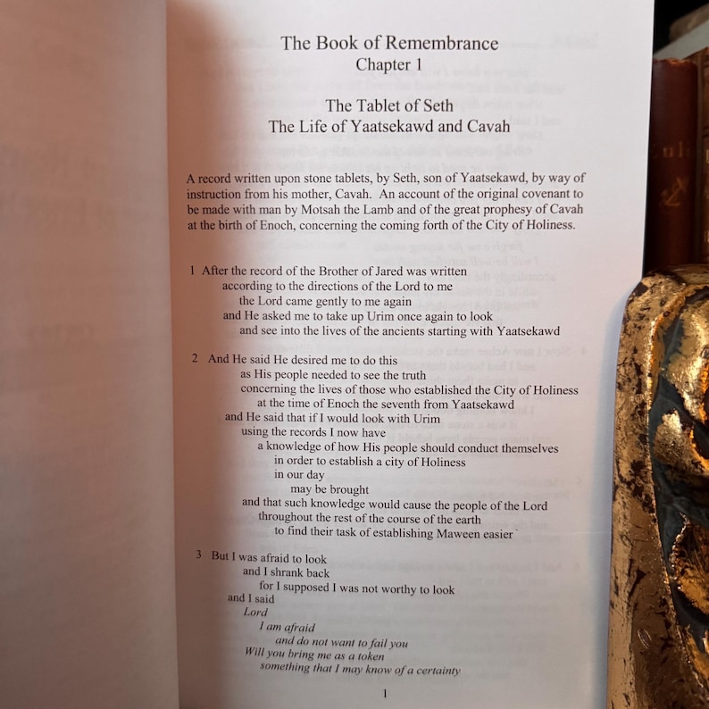 May include: Open book page with text titled "The Book of Remembrance, Chapter 1". The text describes "The Tablet of Seth" and "The Life of Yaatsekawd and Cavah". The page is white with black text. A decorative gold object is on the right.