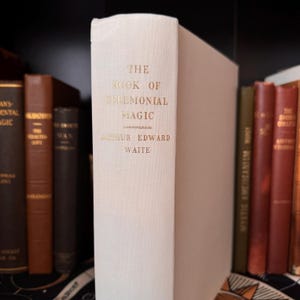 Könnte beinhalten: Ein cremefarbenes Buch mit dem Titel "The Book of Ceremonial Magic" von Arthur Edward Waite. Der Titel und der Name des Autors sind in goldener Schrift. Weitere Bücher sind auf einem dunklen Regal zu sehen.