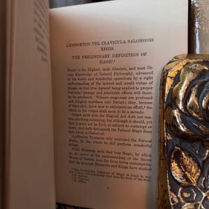 Op de afbeelding: Een open boek met tekst uit "LEMEGETON VEL CLAVICULA SALOMONIS REGIS" en "THE PRELIMINARY DEFINITION OF MAGIC." Het boek heeft een decoratieve goud-zwarte kaft. De pagina's zijn verouderd en vergeeld.