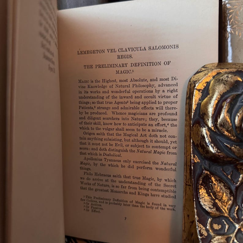 Op de afbeelding: Een open boek met tekst uit "LEMEGETON VEL CLAVICULA SALOMONIS REGIS" en "THE PRELIMINARY DEFINITION OF MAGIC." Het boek heeft een decoratieve goud-zwarte kaft. De pagina's zijn verouderd en vergeeld.
