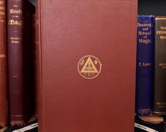 May include: A red hardcover book with gold lettering and a gold symbol on the front cover. The symbol is a triangle with a rope border and an anchor, a star, and a triangle inside. The text on the cover reads "Doctrin and Ritual of Magic" and "E. Levi".