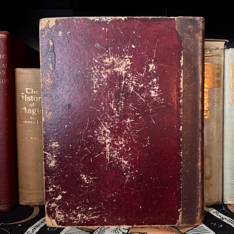 Puede incluir: Un libro antiguo con una cubierta de cuero burdeos desgastada. La cubierta muestra un desgaste significativo con ara&ntilde;azos y &aacute;reas descoloridas. El lomo es de un marr&oacute;n m&aacute;s oscuro. Otros libros son visibles en el fondo, incluido uno con el t&iacute;tulo "The History of Magic".