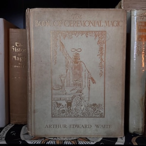 May include: A vintage book titled "The Book of Ceremonial Magic" by Arthur Edward Waite. The cover features a gold illustration of a robed figure with an infinity symbol above their head. The book is light beige with gold lettering.