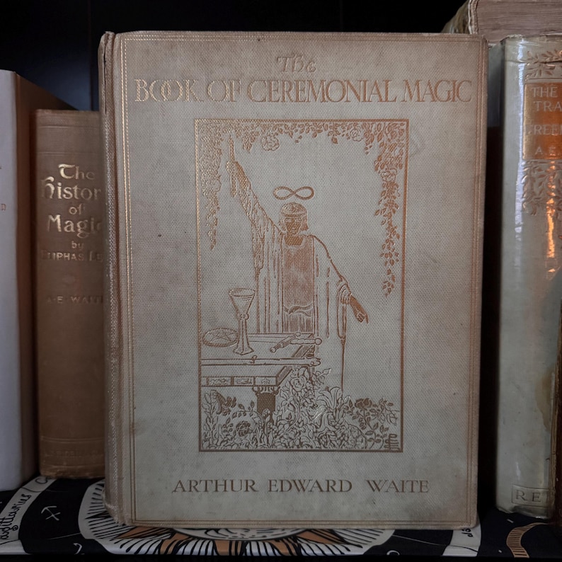 May include: A vintage book titled "The Book of Ceremonial Magic" by Arthur Edward Waite. The cover features a gold illustration of a robed figure with an infinity symbol above their head. The book is light beige with gold lettering.