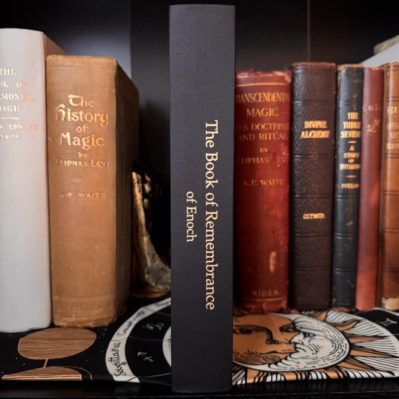 May include: A collection of antique books on a shelf, featuring a black book titled "The Book of Remembrance of Enoch" with gold lettering. Other books have brown and white covers with gold lettering.