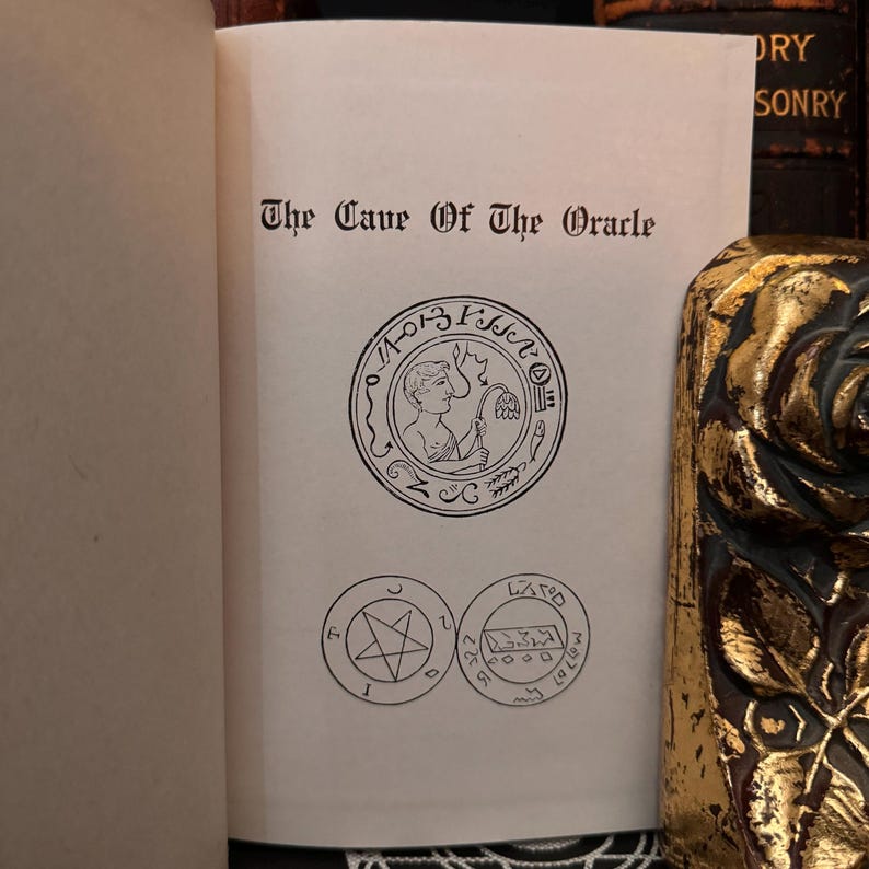 May include: A book titled "The Cave of the Oracle" with a black and white illustration of a seal featuring a person holding a flower. The seal is surrounded by a circle of text.