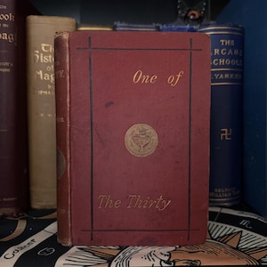 Könnte beinhalten: Ein rotes Hardcover-Buch mit goldenen Lettern und einem goldenen Emblem auf der Vorderseite. Der Titel des Buches lautet "One of The Thirty".