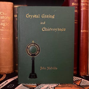 Könnte beinhalten: Ein grünes Hardcover-Buch mit dem Titel "Crystal Gazing and Clairvoyance" von John Melville. Der Buchumschlag zeigt eine goldene und schwarze Illustration eines Kreuzes und eines Kreises mit den Namen "Raphael, Michael, Gabriel, Uriel" darauf.