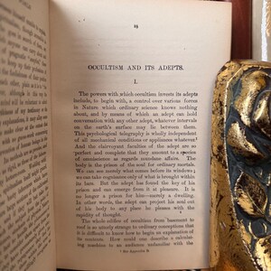 Pu&ograve; includere: Un libro vintage con una pagina ingiallita aperta su un capitolo intitolato "Occultismo e i suoi adepti." Il testo tratta dei poteri degli adepti, tra cui il controllo sulle forze naturali e la comunicazione con altri adepti.
