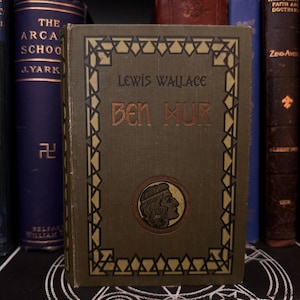 Puede incluir: Un libro verde con un borde dorado y el título "Ben Hur" de Lewis Wallace. La portada del libro presenta una ilustración dorada de la cabeza de una persona de perfil.