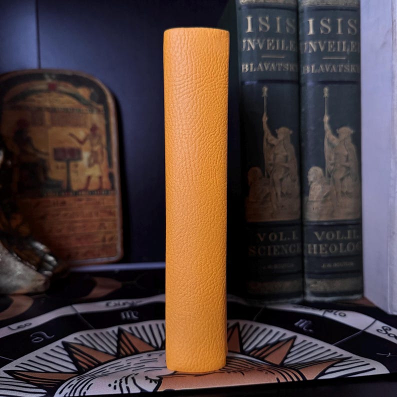 May include: A tall, cylindrical object with a textured, orange-yellow exterior stands upright. Behind it are two dark green books with gold lettering and illustrations. The background includes a decorative piece and a patterned cloth.