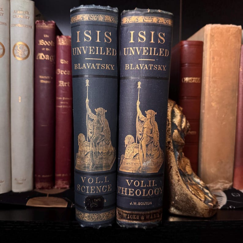 May include: Two antique books titled "Isis Unveiled" by Blavatsky. The dark blue books feature gold lettering and illustrations of a robed figure. Volume I is labeled "Science" and Volume II is labeled "Theology". Other books are visible in the background.