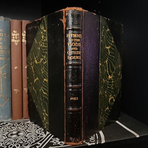 Könnte beinhalten: Antikes Buch mit dem Titel "Hymns to the Gods and Other Poems" mit goldener Schrift auf dem Buchrücken. Das Buch hat einen schwarzen Lederrücken und gemusterte grüne und schwarze Einbände. Weitere Bücher sind im Hintergrund zu sehen.