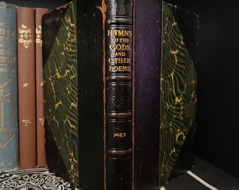 Peut inclure: Livre ancien intitulé "Hymns to the Gods and Other Poems" avec des lettres dorées sur le dos. Le livre a un dos en cuir noir et des couvertures vertes et noires à motifs. D'autres livres sont visibles en arrière-plan.