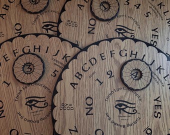 May include: Several wooden talking boards with scalloped edges are arranged together. Each board features the alphabet, numbers, and the words "YES" and "NO." A small, circular pointer with a number is on each board. The boards have a light brown wood grain.