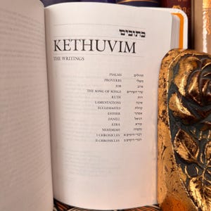May include: An open book with the title "Kethuvim" and the words "The Writings" visible. The book's pages display a list of biblical books, including Psalms, Proverbs, and Job. A decorative gold object is on the right side of the image.