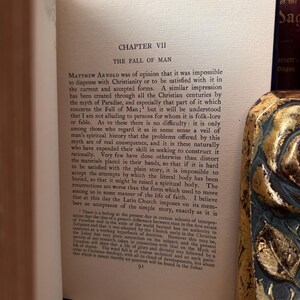 May include: An open book with the title "Chapter VII The Fall of Man" printed on the page. The text is in black ink. A decorative gold and blue book spine is visible on the right. The book appears to be an antique.