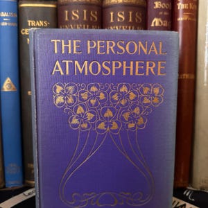Puede incluir: Un libro antiguo titulado "THE PERSONAL ATMOSPHERE" con letras doradas y un dise&ntilde;o floral en una cubierta p&uacute;rpura. Otros libros antiguos son visibles en una estanter&iacute;a.