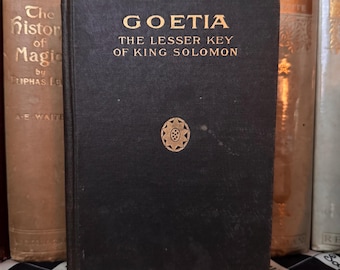 Puede incluir: Un libro antiguo negro titulado "Goetia: The Lesser Key of King Solomon" con letras doradas y un emblema dorado. El libro forma parte de una colección, con otros libros visibles en el fondo, sugiriendo un tema histórico.