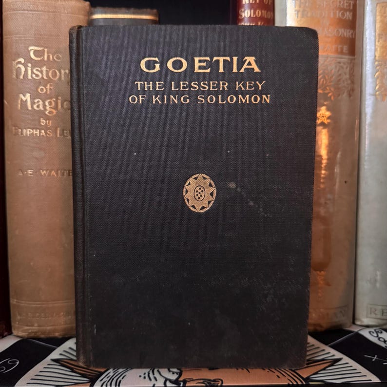 Op de afbeelding: Een zwart, antiek boek met de titel "Goetia: The Lesser Key of King Solomon" met gouden letters en een gouden embleem. Het boek maakt deel uit van een verzameling, met andere boeken op de achtergrond.