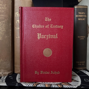 Könnte beinhalten: Ein rotes Buch mit dem Titel "The Chalice of Ecstasy Parzival" mit goldener Schrift und einem kreisförmigen Emblem. Das Buch ist von anderen antiken Büchern mit goldenen und braunen Einbänden umgeben. Der Name des Autors, "By Frater Achad", ist unten aufgedruckt.