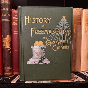 Op de afbeelding: Een groen hardcover boek met de titel "History of Freemasonry and Concordant Orders" met gouden letters en illustraties. Het boek maakt deel uit van een verzameling antieke boeken op een plank. De omslag toont een ster en vrijmetselaars symbolen.