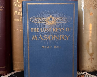 Könnte beinhalten: Ein blaues Buch mit dem Titel "The Lost Keys of Masonry" von Manly Hall. Das Buch hat goldene Buchstaben und ein goldenes Emblem auf dem Einband. Das Buch ist von anderen antiken Büchern umgeben.