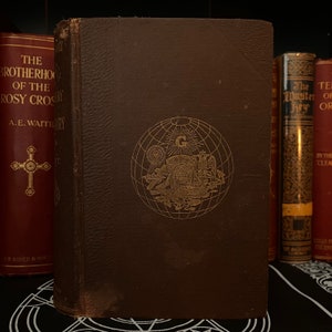 Può includere: Un libro rilegato in pelle marrone con lettere dorate e un'immagine dorata in rilievo di un globo con simboli. Il titolo del libro è "Il Mistero della Rosa-Croce" di A.E. Waite.