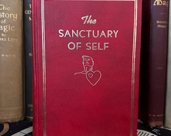 May include: A red hardback book with gold lettering titled "The Sanctuary of Self". The book cover features a line drawing of a person with a heart.