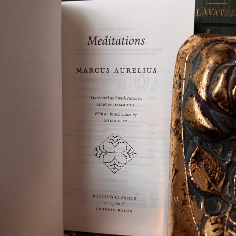 May include: An open book titled "Meditations" by Marcus Aurelius, published by Penguin Classics. The book's pages are white with black text. A decorative, gold-colored object is partially visible on the right side of the image.