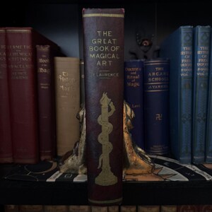 Pu&ograve; includere: Un libro rilegato in bordeaux dal titolo "The Great Book of Magical Art" di E. Laurence. Il libro presenta un'illustrazione dorata di un serpente avvolto attorno a una colonna.