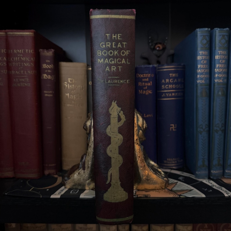 Pu&ograve; includere: Un libro rilegato in bordeaux dal titolo "The Great Book of Magical Art" di E. Laurence. Il libro presenta un'illustrazione dorata di un serpente avvolto attorno a una colonna.